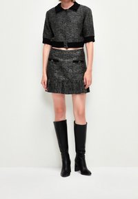 Veste en tweed gris coupée avec une coupe carrée, col noir et accents de boutons, associée à une jupe assortie avec des poches et un ourlet plissé. Bottes noires montantes.