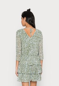 Vestido verde com padrão, com costas em forma de V, mangas até três quartos e volantes em camadas na barra. Feito de um tecido leve e texturizado.