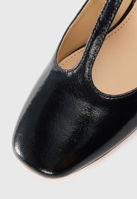 Black patent leather shoe with a square toe. Features cutouts on the sides and a smooth texture. The interior is beige.