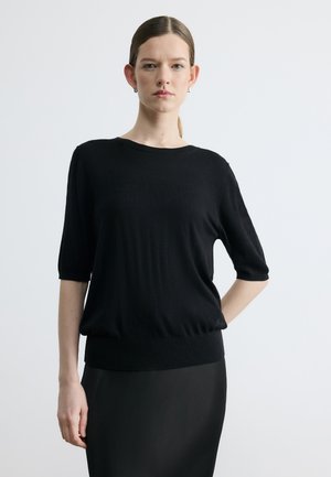 Kvinde med tilbageglattet hår iført en sort kortærmet sweater og sort nederdel, stående med den ene hånd på hoften mod en ensfarvet baggrund.