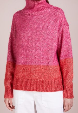 Femme portant un pull à col roulé bloc de couleurs rose et rouge à manches longues et un pantalon blanc, vue des épaules aux hanches.