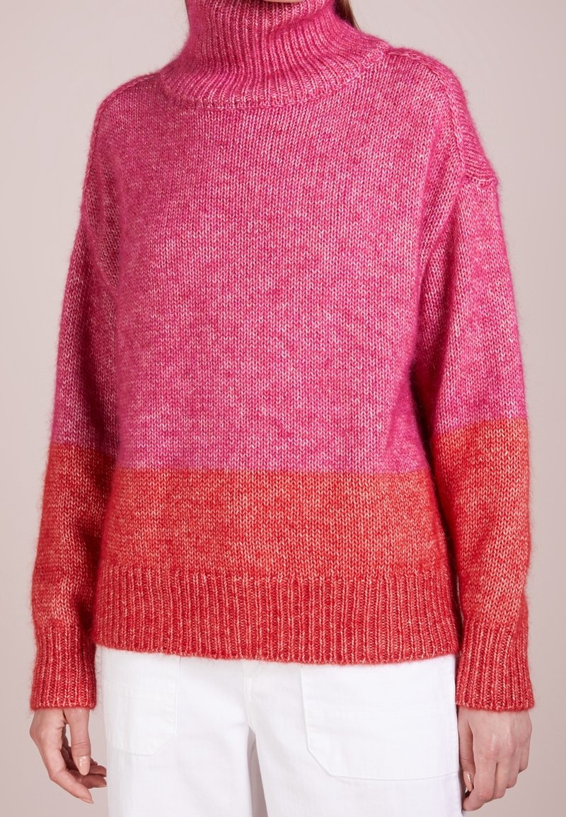 Femme portant un pull à col roulé bloc de couleurs rose et rouge à manches longues et un pantalon blanc, vue des épaules aux hanches.
