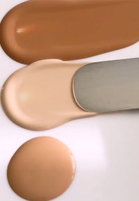 Tre flytende foundation-prøver på en hvit overflate: lys beige, medium beige og rik brun. Jevn tekstur, blank finish.