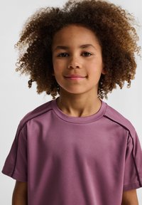 Krótki rękaw, fioletowa koszulka sportowa z teksturowanej tkaniny, okrągły dekolt i detale na szwie ramion, noszona przez dziecko z kręconymi włosami.