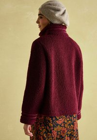 Burgundia fleecest jakk, millel on tekstureeritud viimistlus, kõrge krae ja ribadega aktsendid; koos lillemustrilise seelikuga.