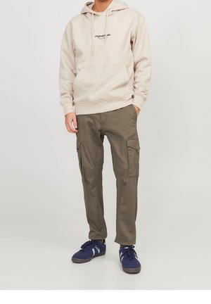 Mann trägt beige Hoodie mit Fronttasche, olivgrüne Cargo-Hose mit Seitentaschen und marineblaue Turnschuhe vor hellem Hintergrund.