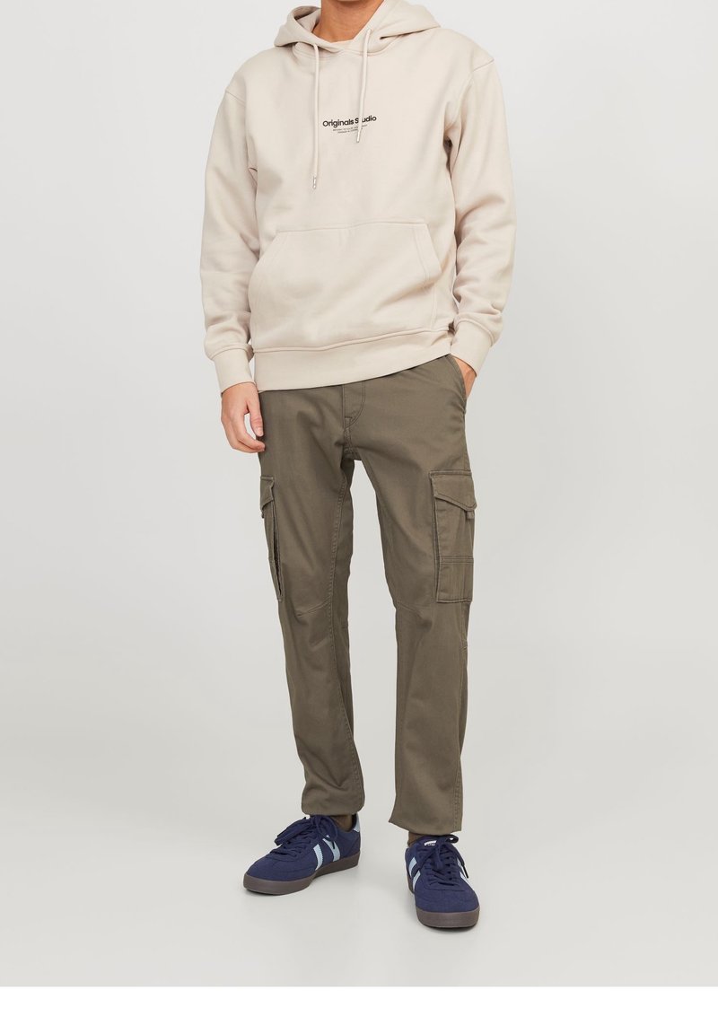Homme portant un sweat à capuche beige avec poche avant, pantalon cargo vert olive avec poches latérales et baskets bleu marine sur fond clair.
