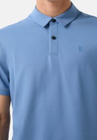 Polo en bleu clair, à manches courtes, en tissu texturé, avec patte de boutonnage à trois boutons, arborant un petit logo en relief de la lettre "B" sur la poitrine.