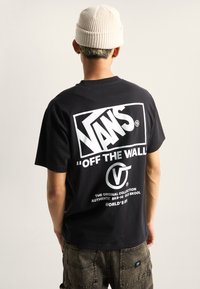 Černé bavlněné tričko s logem "VANS" a grafikou "OFF THE WALL" v bílé barvě na zadní straně. Pod ním jsou viditelné tmavé texturované kalhoty.