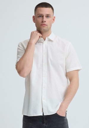 Mężczyzna z krótkimi włosami, ubrany w białą koszulę z krótkim rękawem, prawej ręki dotykającej szyi, lewa ręka w kieszeni, neutralne tło.