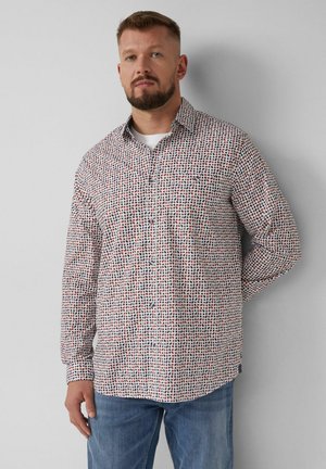 Mężczyzna z brodą w koszuli zapinanej na guziki z wzorem i niebieskich dżinsach, stojący z jedną ręką za plecami na tle gładkiej ściany.