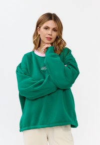 Felpa verde oversize con colletto rotondo rosa e un piccolo logo bianco. Il tessuto appare morbido con una texture liscia e una vestibilità comoda.