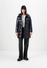 Vrouw staat rechtop in een marineblauwe gewatteerde jas met capuchon, witte shirt, zwarte wijde broek en zwarte chunky schoenen tegen een witte achtergrond.