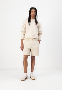 Beige sweatshirt with embroidered detail, paired with light beige shorts. Model wears white shoes and light gray socks. Simple, relaxed fit.