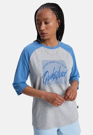 Młoda osoba z warkoczami, ubrana w szaro-niebieską koszulkę Quiksilver, patrząca w dół, z rękami splecionymi z przodu, neutralne tło.