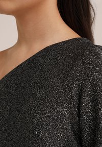 Asymmetrisk svart topp med en texturerad, metallisk finish. Har en slät halsringning och puffärmsdetalj på högra sidan.