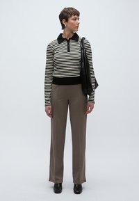 Polo de manga comprida às riscas pretas e brancas com gola preta, combinado com calças castanhas de pernas largas e sapatos pretos, a carregar uma mala preta.