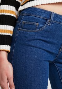 Jeans de color azul oscuro con una textura suave, que presentan un bolsillo trasero y detalles de costura sutiles. Combinados con un suéter a rayas en negro, blanco y amarillo.