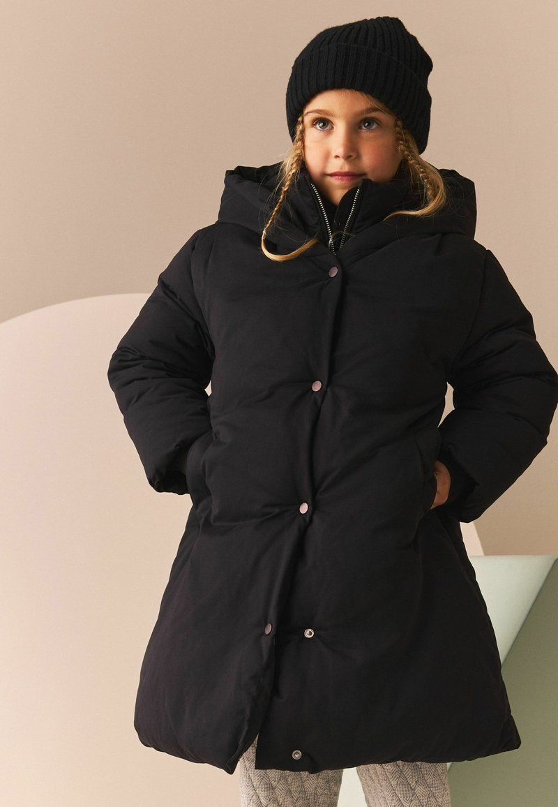 Geacă puf cu капюшon neagră, cu nasturi lucioși și textură matlasată. Purtați peste un top negru și pantaloni cu model. Beanie tricotat negru.