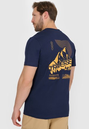 Mężczyzna w granatowej koszulce z żółtą grafiką góry i napisem „TRAVERSE est. 1983”, beżowe szorty, patrzy przez ramię.