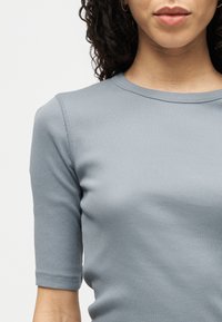 Kvinne iført en ribbestrikket, kortermet, lys grå skjorte med rund hals, vist fra skuldrene til øvre del av brystet og leppene.