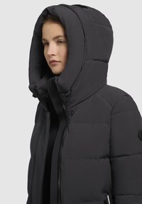 La giacca a vento nera presenta un grande cappuccio, chiusura con zip e bottoni a pressione. Il tessuto è liscio con una texture trapuntata e dettagli con logo.