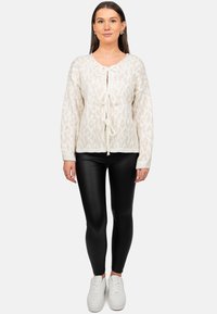 Cardigan lavorato a maglia color crema con motivo leopardato, chiusura frontale con lacci, polsini a coste e finitura testurizzata, abbinato a leggings neri e sneakers bianche.