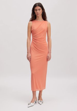 Frau mit langen dunklen Haaren, die ein ärmelloses, gerafftes korallenfarbenes Midikleid und weiße spitze Slingback-High Heels vor einem einfarbigen Hintergrund trägt.