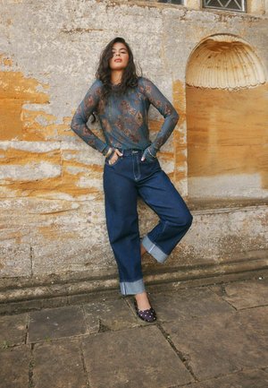 Frau lehnt an einer verwitterten Steinmauer, trägt ein durchsichtiges, gemustertes Langarmshirt, weit geschnittene, umgekrempelte Jeans und gepunktete Ballerinas.