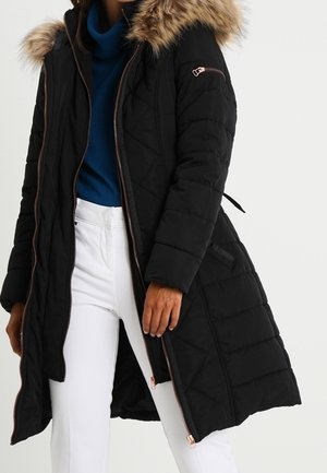 Femme portant un long manteau noir matelassé avec une capuche en fourrure, un pull à col roulé bleu et un pantalon blanc, debout avec une main le long du corps.