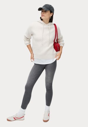 Witte geribbelde hoodie, grijze legging, rode handtas, witte schoenen met rode accenten en een donkere pet. Model staat met één hand op de heup.