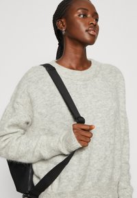 Šviesiai pilkas tekstūruotas megztinis su laisvu siluetu ir vyniojamaisiais kaklo kraštais; dėvimas su juoda petnešų rankine, akcentuojančia mezgimo rankogalius ir siūlių detales.