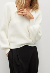 Biały sweter z prążkowanym splotem, z dekoltem w serek i luźnym krojem. Długie rękawy mają lekki bufkę na ramieniu. W zestawie z czarnymi spodniami.