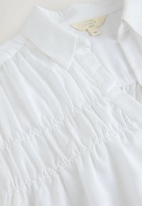 Biała lniana koszula z kołnierzykiem, z marszczeniem na przodzie, miękka w dotyku i o prostych elementach designu.