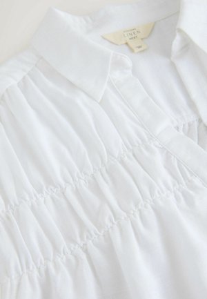 Weißes Leinenhemd mit Kragen, das am Oberteil geraffte Details, eine weiche Textur und schlichte Designelemente aufweist.