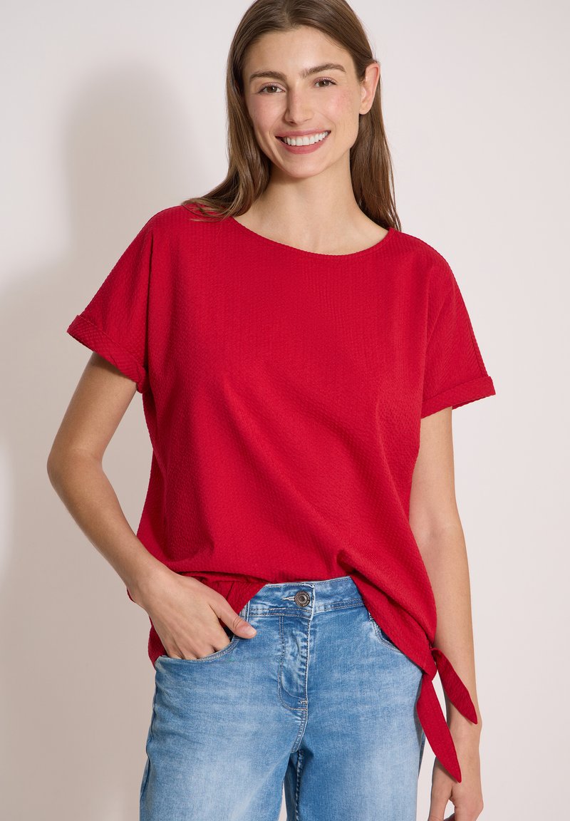 Femme souriante portant un haut rouge à manches courtes avec des liens sur les côtés et un jean bleu clair, debout devant un fond clair uni.