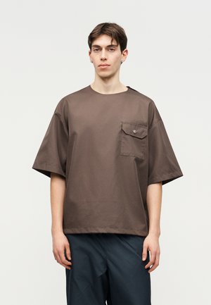 Fiatal férfi rövid sötét hajjal, laza barna rövid ujjú ingben, mellzsebbel, sötétkék nadrágban, sima háttér előtt állva.