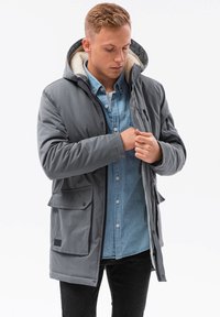 Grå täckjacka med huva, med en mjuk fuskpälsfoder, dragkedja och tryckknappar framtill, samt stora sidofickor. Bärs över en jeansskjorta.