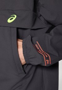 Schwarze Jacke mit strukturierter Oberfläche. Verfügt über ein grünes Logo und roten "ASICS TRACK CLUB"-Text auf dem Ärmel. Elastische Bündchen, sichtbare Reißverschlusstasche.
