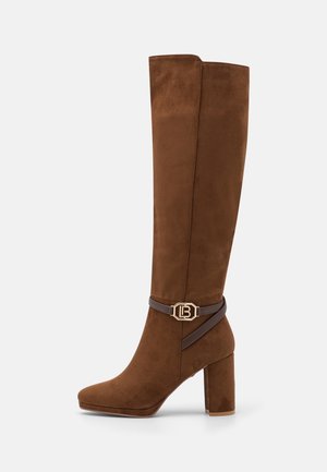 Botte haute en daim marron avec talon bloc et bride décorative marron foncé avec boucle dorée autour de la cheville.