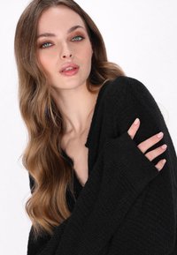Schwarzer Strickpullover mit V-Ausschnitt, lockerer Passform und langen Ärmeln. Das Model posiert mit einem verschränkten Arm und zeigt die Textur und das Design.