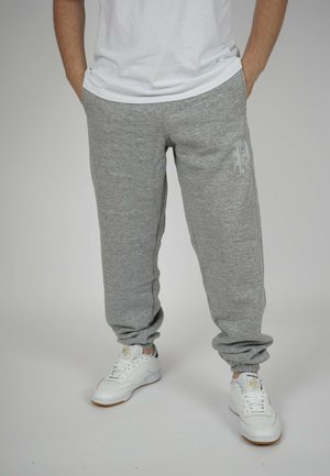 Graue Baumwoll-Sweatpants mit elastischen Bündchen und einem dezenten Logo auf dem Oberschenkel, kombiniert mit weißen Sneakers, die eine Gummisohle und strukturierte Details aufweisen.