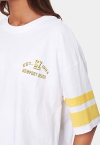 Žena v bílém oversized tričku s žlutými pruhy na rukávech a žlutým textem "EST. SZ 1984 NEWPORT BEACH" na hrudi.