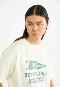 Camiseta de algodón color crema con un gráfico verde que presenta "NEW BALANCE ATHLETICS" y un diseño de estandarte. Manga corta, cuello redondo.