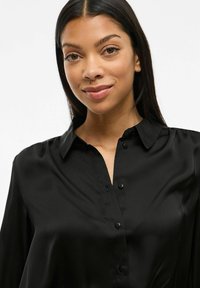 Camicia nera in satin con collo e maniche lunghe, caratterizzata da una texture liscia e semplici bottoni neri lungo la parte frontale.
