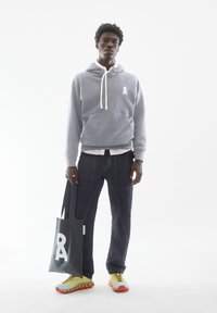 Junger Mann trägt grauen Hoodie, dunkle Hose, gelbe und orangefarbene Sneakers, hält eine schwarze Tragetasche mit weißem Logo vor weißem Hintergrund.