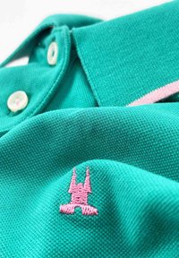 Polo turquoise avec un col, des boutons blancs, et un logo brodé rose avec un design distinctif sur le devant. Texture douce.