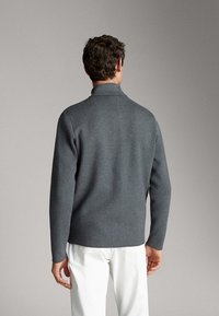Maglione grigio a maglia con colletto, design aderente e tessuto testurizzato, indossato sopra pantaloni bianchi, visto da dietro.
