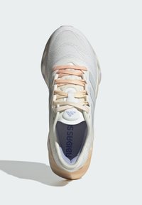 Scarpa sportiva bianca con superiore testurizzato, lacci rosa e crema, e suola beige chiaro. Presenta un logo sulla linguetta e dettagli a strisce.