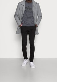 Cappotto grigio in lana, felpa nera con logo, pantaloni neri slim-fit e sneakers bianche. Il cappotto presenta un colletto a punte e chiusura con bottoni.